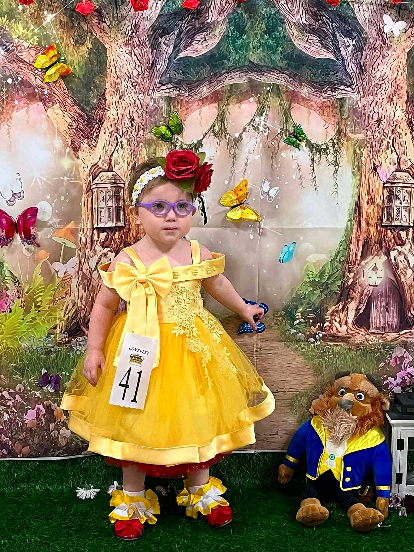 Amelia joined the competition — help win amazing prizes! child, girl, toddler, yellow_dress, bow, headband, rose, glasses, butterflies, backdrop, stuffed_animal, plush_toy, pageant, contest_number, red_shoes, grass, fairy_tale, portrait, indoor, costume