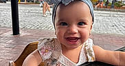 Amari is registered to the contest to win money with this photo: baby, child, smiling, headband, bow, hand, table, outdoor, daylight, tree, street, people, blurred_background, happy, portrait, cute, clothing, earrings, highchair, cobblestone