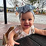 baby, child, smiling, headband, bow, hand, table, outdoor, daylight, tree, street, people, blurred_background, happy, portrait, cute, clothing, earrings, highchair, cobblestone