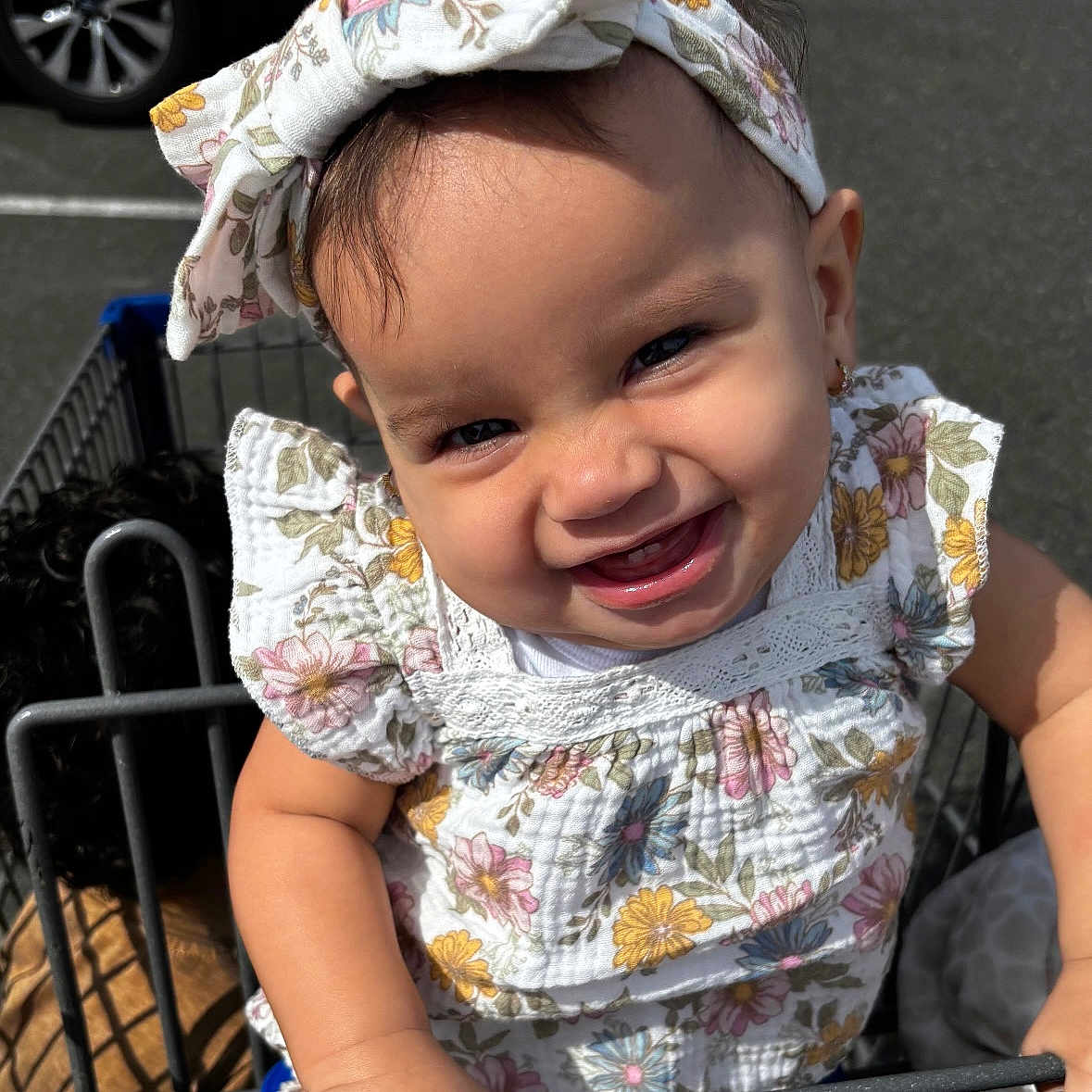 Amari is registered to the contest to win money with this photo: baby, child, shopping_cart, headband, smile, floral_pattern, outdoor, sunlight, cute, happy, toddler, clothing, person, holding, face, hand, playful, parked_car, parking_lot, expression