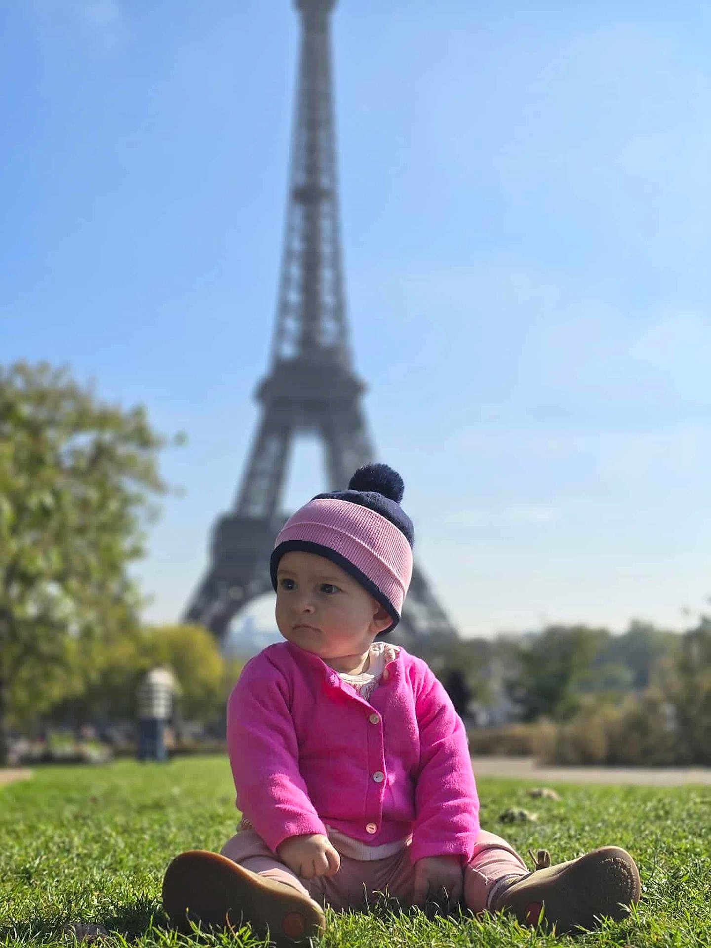 Lynda-Rose participe au concours pour gagner de l'argent avec cette photo : toddler, child, grass, hat, pink_sweater, shoes, outdoor, daylight, eiffel_tower, landmark, sky, trees, park, sitting, portrait, person, nature, casual_clothing, cute, urban