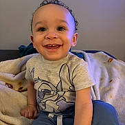 Jonah is registered to the contest to win money with this photo: baby_photos, blanket, child, clothing, comfortable, cozy, curly_hair, cute, face, happy, home, indoor, pants, person, portrait, sitting, smiling, toddler, tshirt, young_child