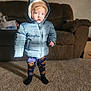 child, toddler, jacket, hat, carpet, couch, indoor, person, face, standing, blue_jacket, brown_couch, wall_art, photograph, cozy, warm_clothing, pants, socks, curious_expression, home