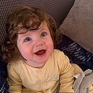 Evan is registered to the contest to win money with this photo: baby, child, curly_hair, blue_eyes, smiling, yellow_onesie, plush_toy, elephant_toy, cushion, soft_texture, indoors, happy, cute, portrait, infant, seated, cozy, young_child, expression, comfort