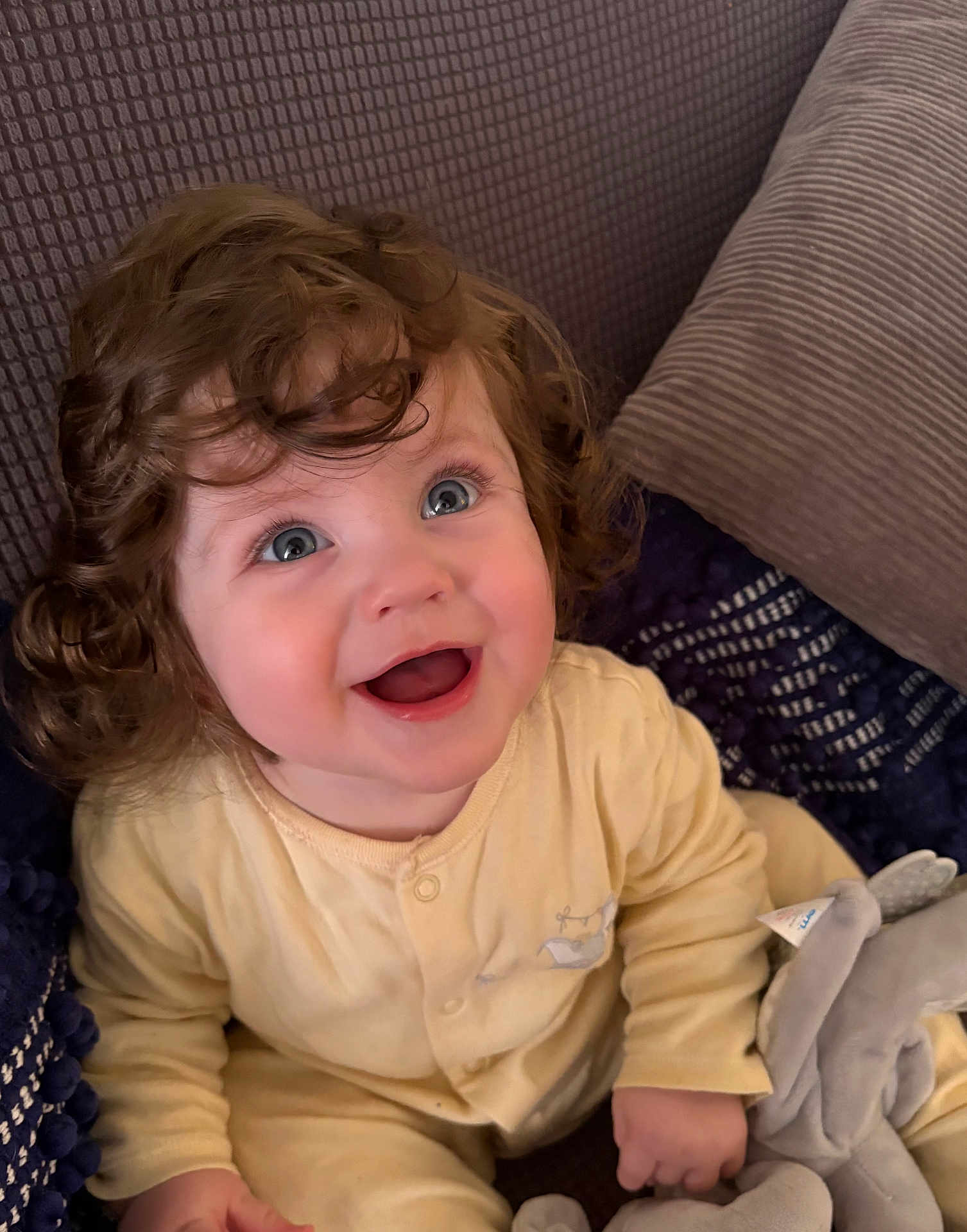 Evan is registered to the contest to win money with this photo: baby, child, curly_hair, blue_eyes, smiling, yellow_onesie, plush_toy, elephant_toy, cushion, soft_texture, indoors, happy, cute, portrait, infant, seated, cozy, young_child, expression, comfort