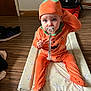 baby, child, pumpkin_costume, pacifier, mat, zebra_pattern, wooden_floor, indoor, curious, orange_clothing, headwear, footwear, vacuum_cleaner, toy, person, flooring, costume, sitting, expression, home