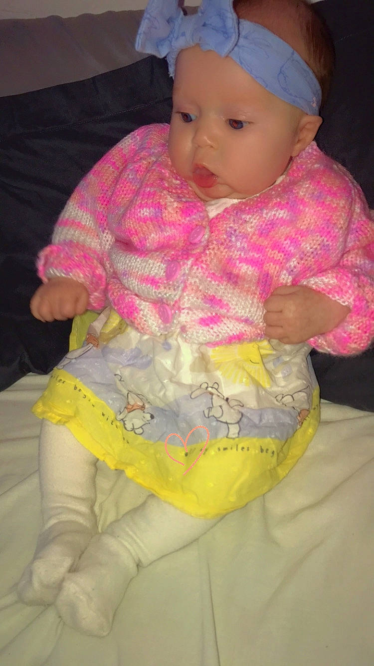 accessories, baby, blanket, bonnet, cap, clothing, coat, couch, dress, face, furniture, hat, head, headband, knitwear, pants, person, sitting, sweater, toy