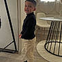 Tiago participe au concours pour gagner de l'argent avec cette photo : child, boy, indoor, standing, smiling, black_turtleneck, beige_pants, sneakers, table, chair, wooden_floor, window, radiator, rug, modern_furniture, home_interior, curtains, playful, portrait, casual_clothing