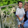 child, boy, smiling, white_tshirt, shorts, sneakers, wristband, outdoor, railing, greenery, daylight, person, casual_clothing, standing, happy, portrait, blurred_background, metal_railing, nature, people