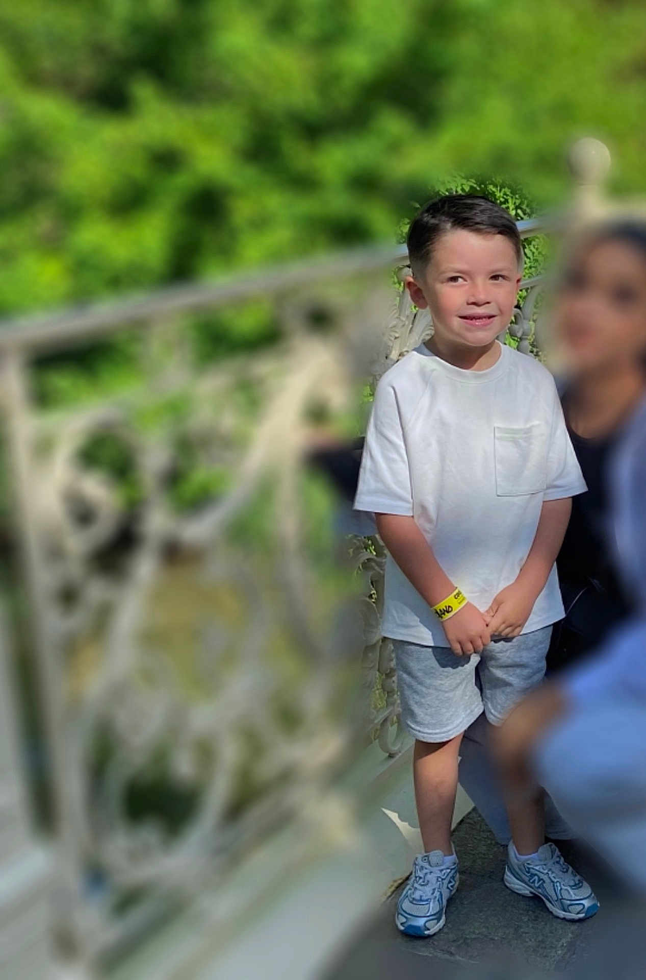 Tiago a rejoint le concours — aidez-le/la à gagner de superbes lots ! child, boy, smiling, white_tshirt, shorts, sneakers, wristband, outdoor, railing, greenery, daylight, person, casual_clothing, standing, happy, portrait, blurred_background, metal_railing, nature, people