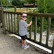 Tiago participe au concours pour gagner de l'argent avec cette photo : child, boy, smiling, sunglasses, striped_shirt, black_shorts, gray_sneakers, wooden_fence, outdoor, nature, trees, river, water, pathway, daylight, greenery, happy, casual_clothing, portrait, park