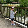 child, boy, smiling, sunglasses, striped_shirt, black_shorts, gray_sneakers, wooden_fence, outdoor, nature, trees, river, water, pathway, daylight, greenery, happy, casual_clothing, portrait, park