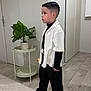 Tiago participe au concours pour gagner de l'argent avec cette photo : child, boy, indoor, plant, potted_plant, table, flooring, clothing, sneakers, black_pants, white_shirt, hairstyle, modern_interior, side_table, neutral_colors, standing, serious_expression, floor_rug, door, wall