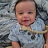 animal_print, baby, bed, blanket, brown_hair, child, close_up, clothing, cozy, eyes, face, happy, indoor, infant, photography, portrait, sitting, smiling, teething_necklace, toy