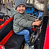 amusement_park, beanie, candid, car, carousel, child, family_fun, happy, jeans, metal_rail, outdoor, playful, portrait, puffy_jacket, red_car, ride, seat, smiling, sneakers, steering_wheel