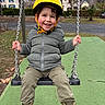 chains, child, grass, green_jacket, happy, helmet, houses, mountains, outdoors, park, play, playground, portrait, puffer_jacket, rubber_boots, smiling_child, swing, swing_seat, winter_season, yellow_helmet