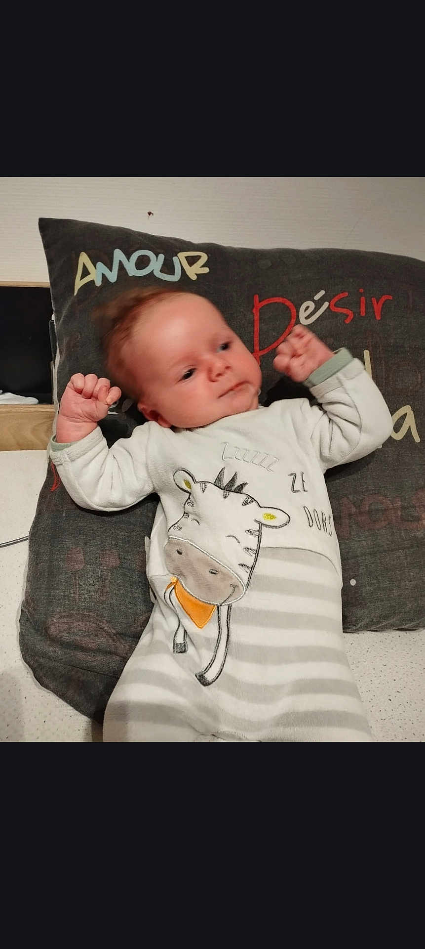 Tylio a rejoint le concours — aidez-le/la à gagner de superbes lots ! baby, child, infant, onesie, striped_clothing, giraffe_design, pillow, text_on_pillow, bed, sleepwear, relaxed, indoor, cute, human, face, hands, lying_down, cozy, soft_lighting, portrait
