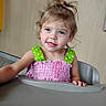 child, toddler, blue_eyes, smile, high_chair, pink_dress, green_straps, polka_dots, curly_hair, ponytail, indoor, cute, person, baby, face, happy, skin, small_hand, seat, portrait
