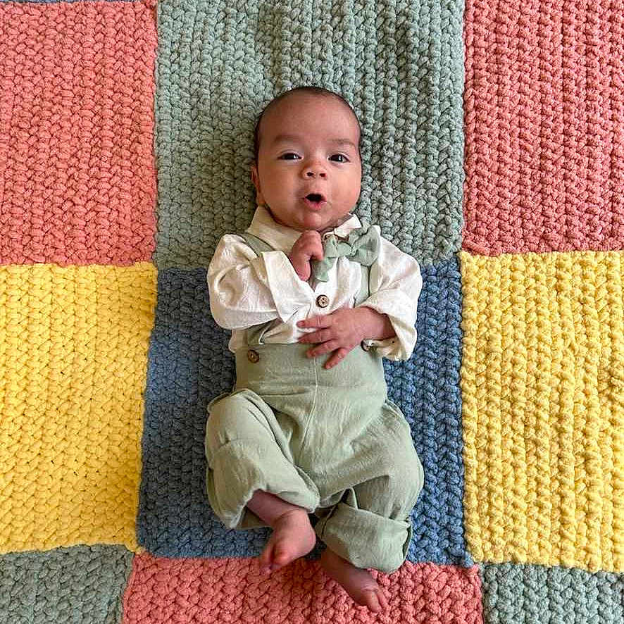 Baby Brent is registered to the contest to win money with this photo: baby, barefoot, bow_tie, child, colorful, cozy, cute, expression, fabric_texture, green_overalls, hand_near_mouth, indoors, infant, knitted_blanket, lying_down, patchwork, playful, portrait, soft_lighting, white_shirt