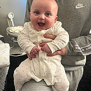 Niamh joined the competition — help win amazing prizes! adult_hand, baby, blanket, child, couch, cozy, cute, expression, face, furniture, grey_clothing, happy, heart_pattern, holding, indoor, infant, nike_logo, onesie, person, smiling