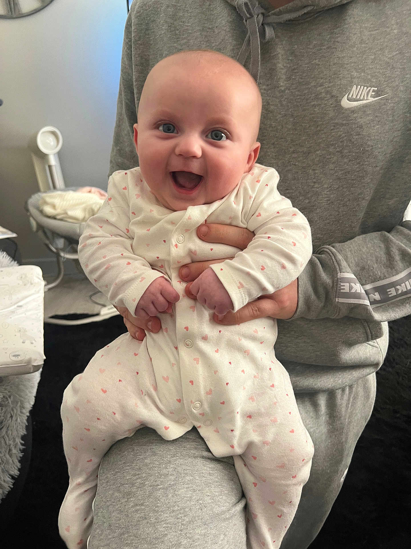 Niamh joined the competition — help win amazing prizes! adult_hand, baby, blanket, child, couch, cozy, cute, expression, face, furniture, grey_clothing, happy, heart_pattern, holding, indoor, infant, nike_logo, onesie, person, smiling