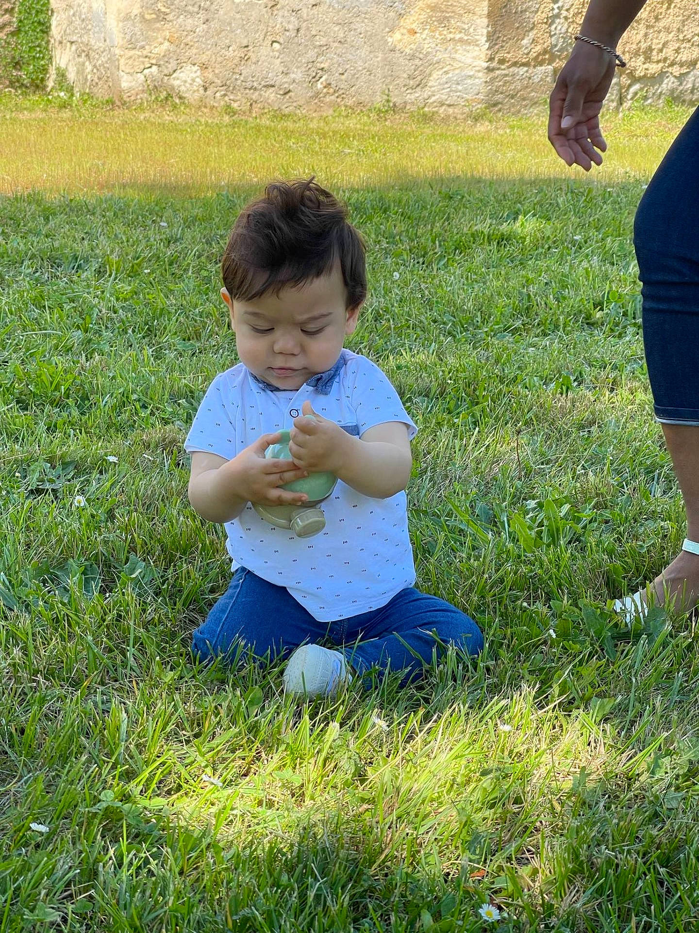 Ahyann-Mathïs a rejoint le concours — aidez-le/la à gagner de superbes lots ! bottle, casual_clothing, child, concentration, curly_hair, daylight, grass, green, hand, jeans, nature, outdoor, person, playing, short_sleeve, sitting, stone_wall, sunlight, toddler, white_shirt
