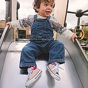 Ahyann-Mathïs a rejoint le concours — aidez-le/la à gagner de superbes lots ! toddler, child, playground, slide, denim_overalls, sweater, curly_hair, smiling, outdoor, fun, shoes, socks, metal_slide, happy, leisure, daytime, park, person, activity, casual_clothing