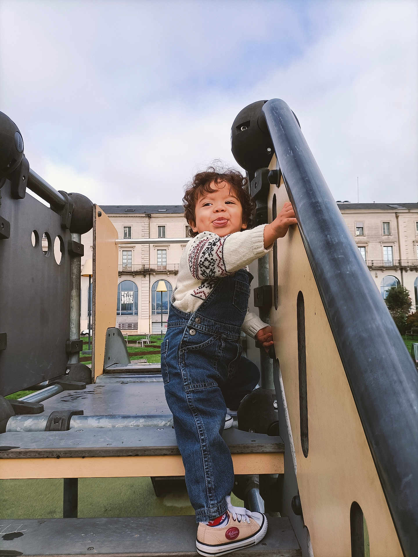 Ahyann-Mathïs participe au concours pour gagner de l'argent avec cette photo : child, toddler, playground, climbing, denim_overalls, sneakers, curly_hair, tongue_out, outdoor, urban, steps, metal, wood, building, sky, cloudy, fun, play, cute, person