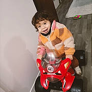 Ahyann-Mathïs participe au concours pour gagner de l'argent avec cette photo : child, colorful, curtain, floor, footwear, furniture, happy, helmet_absent, indoor, jacket, living_room, person, playful, red, smiling, toddler, toy_motorcycle, vehicle, wood_floor, young_child