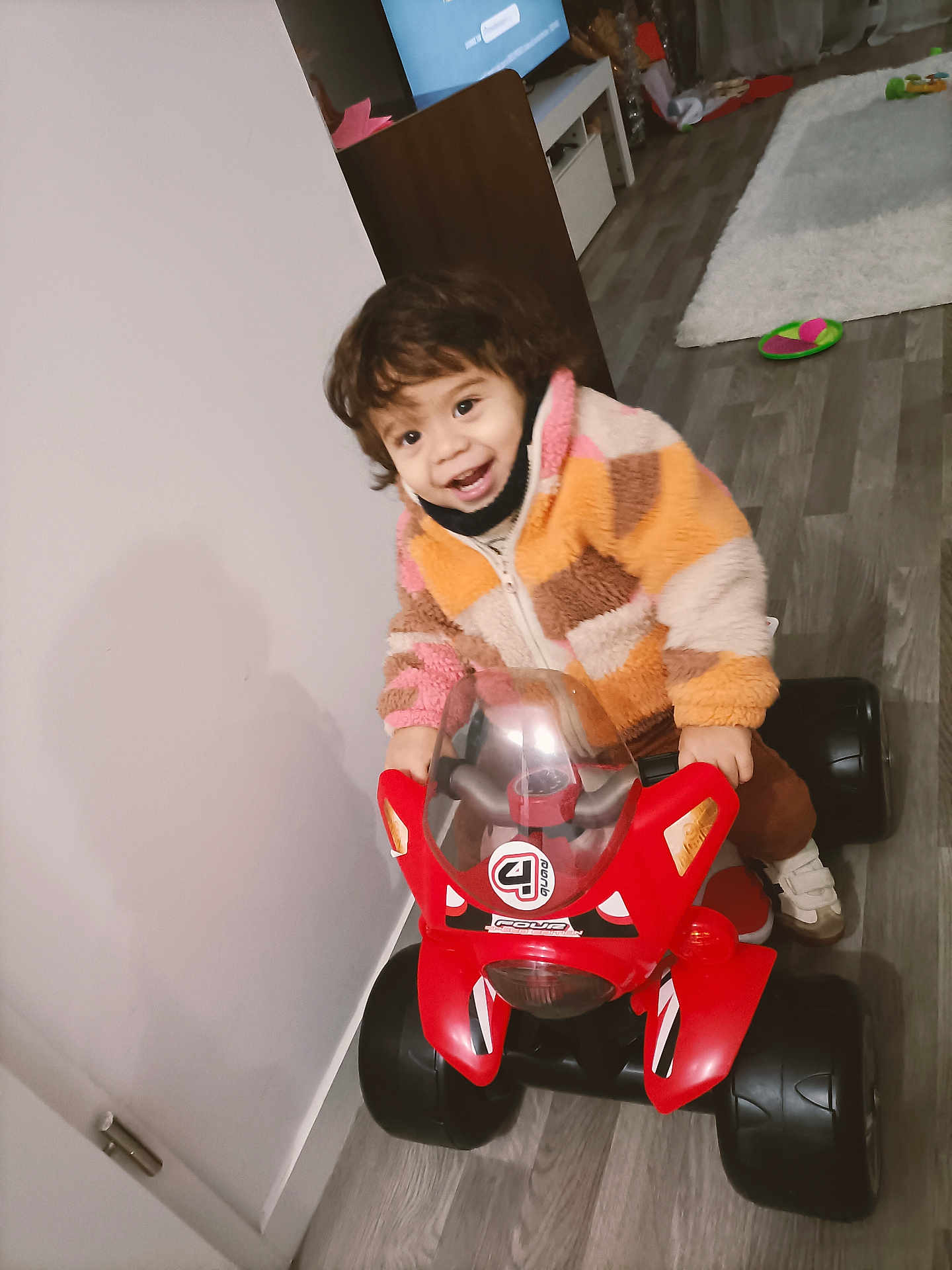 Ahyann-Mathïs participe au concours pour gagner de l'argent avec cette photo : child, colorful, curtain, floor, footwear, furniture, happy, helmet_absent, indoor, jacket, living_room, person, playful, red, smiling, toddler, toy_motorcycle, vehicle, wood_floor, young_child