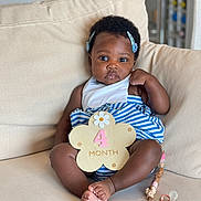 Naïlah participe au concours pour gagner de l'argent avec cette photo : baby, infant, child, couch, sofa, clothing, bib, hair_clip, pacifier, beaded_clip, flower_sign, striped_outfit, skin, foot, hand, sitting, indoor, cute, portrait, young_child