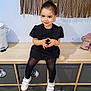 toddler, child, black_dress, white_shoes, sitting, bench, cubicles, storage_bins, wood_floor, wooden_furniture, indoor, smiling, happy, portrait, cute, face, hands, hair_bun, wall_decor, plastic_trash_can