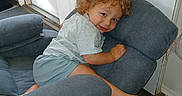 Damien joined the competition — help win amazing prizes! child, toddler, curly_hair, smiling, portrait, sitting, lounging, couch, sofa, cushions, indoor, home, door, window, wood_floor, barefoot, shorts, shirt, playful, face