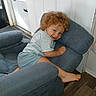 child, toddler, curly_hair, smiling, portrait, sitting, lounging, couch, sofa, cushions, indoor, home, door, window, wood_floor, barefoot, shorts, shirt, playful, face