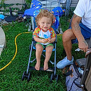 Damien is registered to the contest to win money with this photo: child, toddler, curly_hair, smiling, stroller, outdoor, garden, grass, garden_hose, patio, adult, family, colorful_clothing, shorts, snack, seated, portrait, backyard, plants, daylight