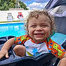 Damien is registered to the contest to win money with this photo: toddler, child, smiling, curly_hair, pool, swimming_pool, poolside, outdoor, sunny, water, summer, swimwear, float, seated, portrait, happy, teeth, blue_sky, backyard, fence