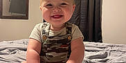 Tucker is registered to the contest to win money with this photo: baby, infant, smile, camo_overalls, bed, bedroom, mirror, curtains, happy, child, person, feet, hands, toes, short_hair, indoor, light, cute, sitting, portrait