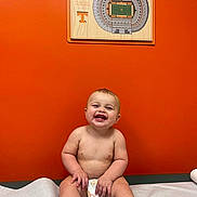 Tucker joined the competition — help win amazing prizes! baby, child, diaper, smiling, sitting, orange_wall, medical_table, indoor, happy, cute, face, skin, barefoot, toes, head, person, room, artwork, stadium, photography