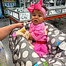 baby, child, pink_clothing, headband, bow, baby_carrier, polka_dots, shopping_cart, store, display_case, curly_hair, hand, toddler, indoor, retail, person, cute, fashion, accessory, shopping