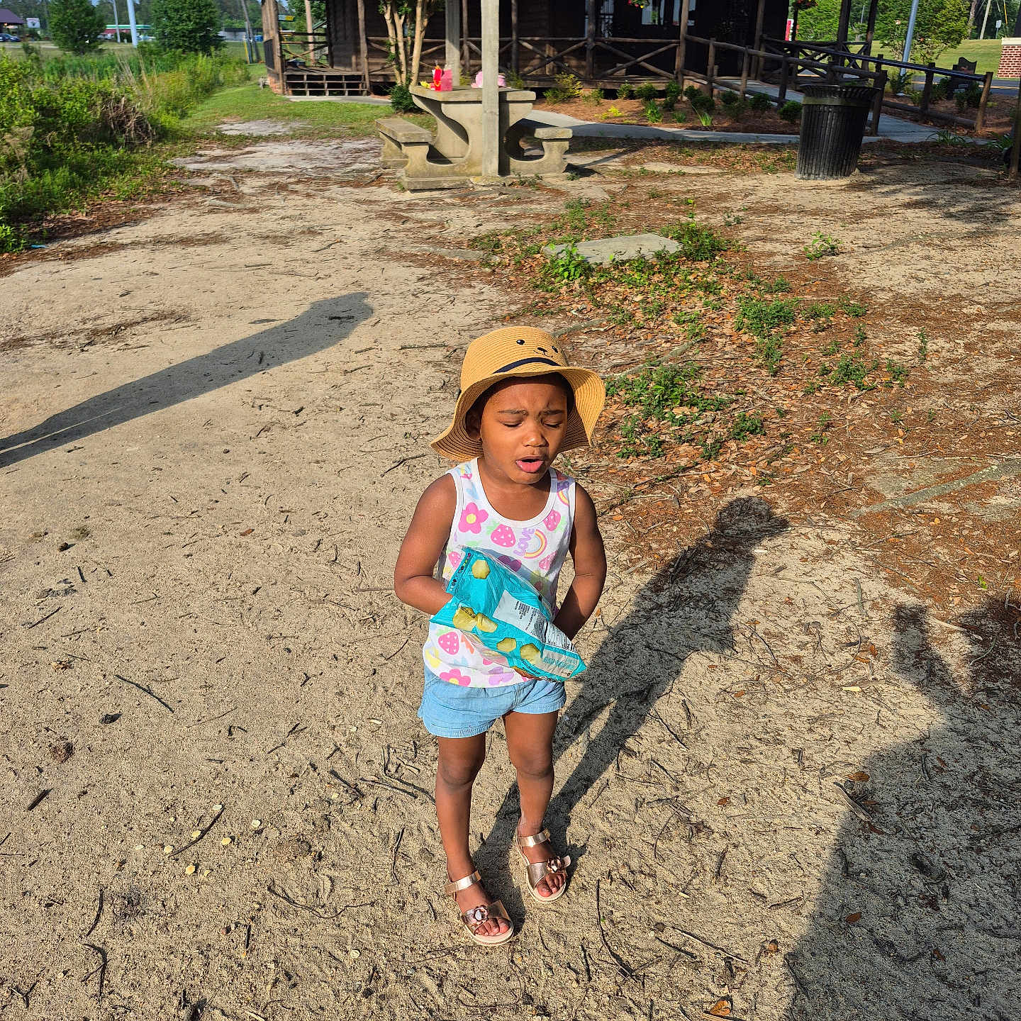 Samantha.lawrence is registered to the contest to win money with this photo: bench, building, casual_clothing, child, daytime, girl, grass, hat, nature, outdoor, sand, shadow, shorts, sidewalk, snack, summer, sunny, tank_top, tree, wooden