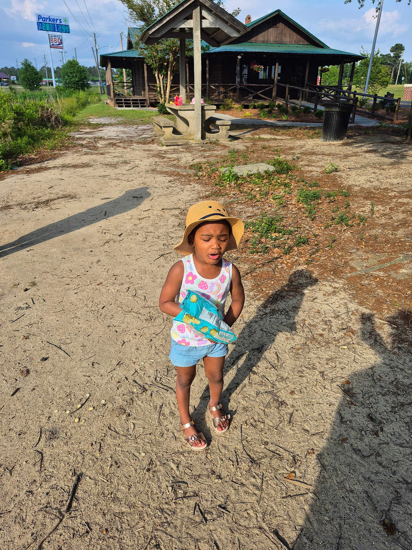 Samantha.lawrence is registered to the contest to win money with this photo: bench, building, casual_clothing, child, daytime, girl, grass, hat, nature, outdoor, sand, shadow, shorts, sidewalk, snack, summer, sunny, tank_top, tree, wooden