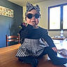 baby, child, table, sunglasses, headband, houndstooth, black_clothing, indoor, window, chair, wooden_table, person, cute, fashion, infant, hand_in_mouth, smiling, home, plant, decor