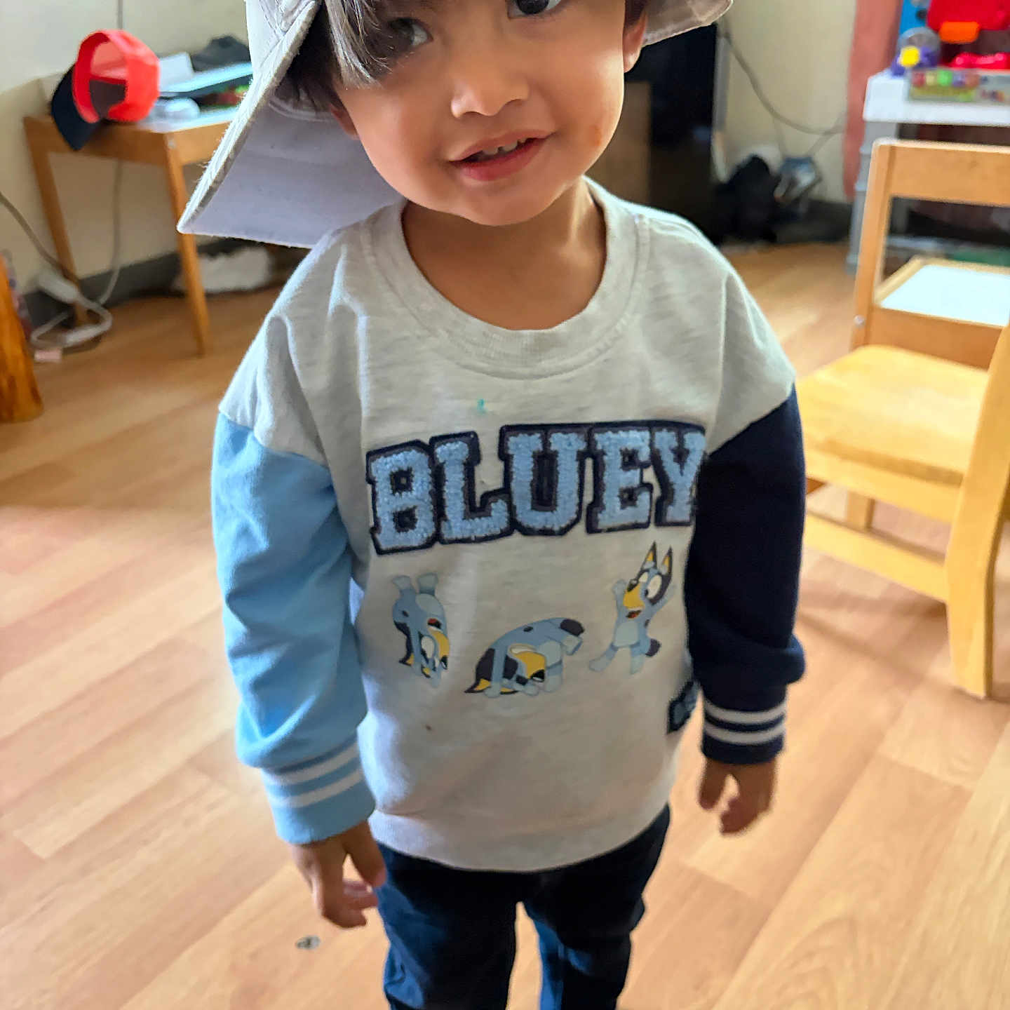 Alessio joined the competition — help win amazing prizes! child, toddler, portrait, smiling, indoors, playroom, hat, floppy_hat, bluey_shirt, long_sleeves, pants, socks, shoes, wooden_floor, chair, table, toys, home, standing, face