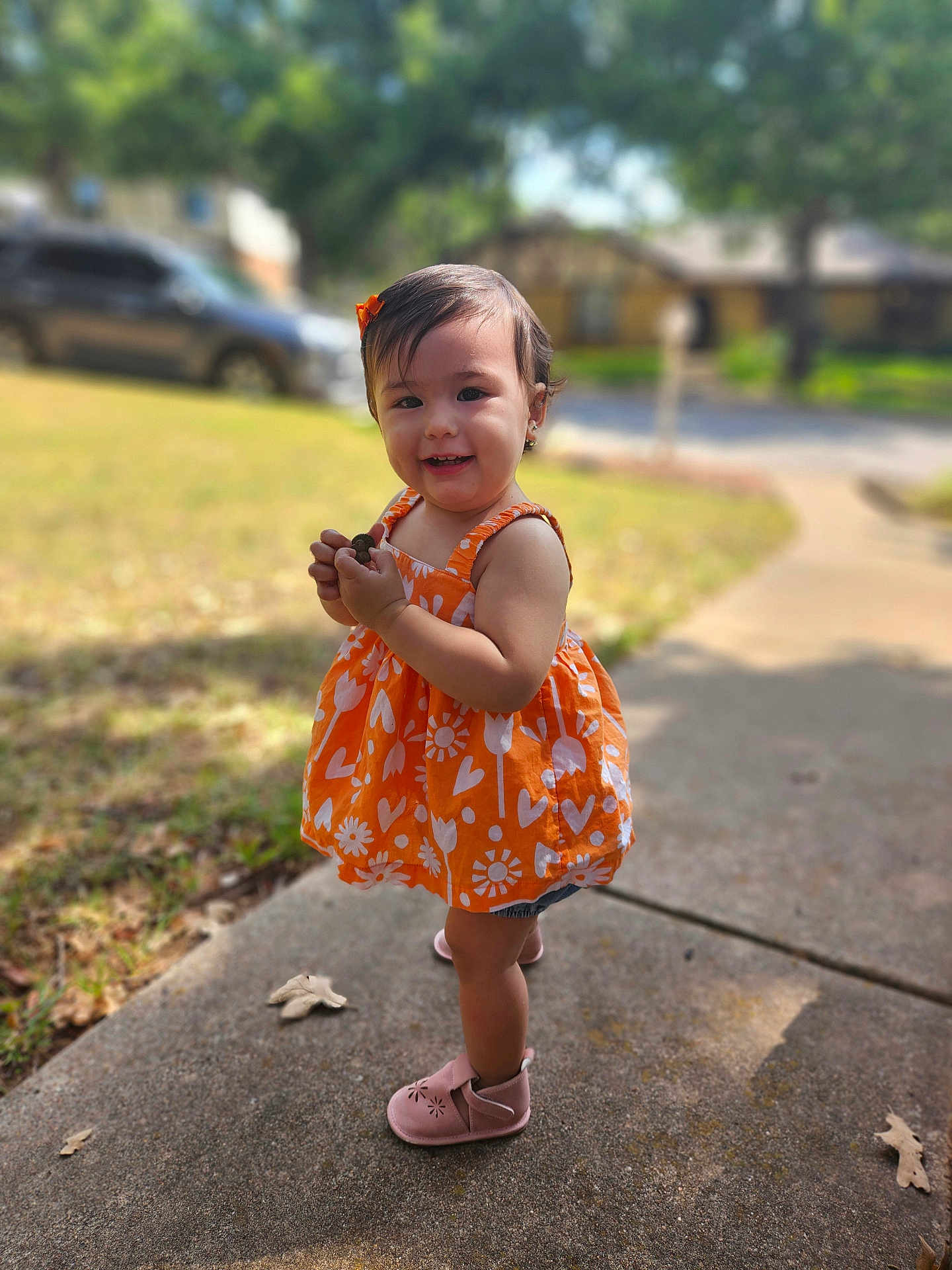 Everly joined the competition — help win amazing prizes! toddler, child, orange_dress, smiling, outdoor, sidewalk, grass, trees, sunlight, pink_shoes, happy, portrait, young_child, person, nature, daylight, flower_pattern, earrings, hair_bow, blurred_background