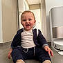 baby, child, smiling, sitting, floor, indoor, clothing, socks, jacket, white_shirt, hallway, door, air_purifier, robot_vacuum, tile_floor, happy, person, home, infant, cute