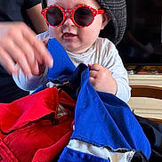 Mayden participe au concours pour gagner de l'argent avec cette photo : baby, child, red_sunglasses, hat, blue_fabric, red_fabric, hand, wooden_table, clock, wood_paneling, indoor, person, playing, table, furniture, face, glasses, adult_hand, television, casual_clothing