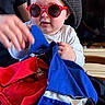 baby, child, red_sunglasses, hat, blue_fabric, red_fabric, hand, wooden_table, clock, wood_paneling, indoor, person, playing, table, furniture, face, glasses, adult_hand, television, casual_clothing