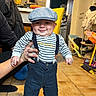 baby, child, smile, striped_shirt, suspenders, cap, hand, floor, tile_floor, indoor, kitchen, chair, bottle, person, clothing, footwear, adult_hand, cabinet, broom, toy
