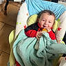 baby, blanket, bottle, child, smiling, indoor, baby_chair, floor, toy, tricycle, soccer_ball, red_clothing, blue_blanket, happy, person, infant, seat, tiled_floor, cute, cozy