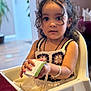 Anand joined the competition — help win amazing prizes! toddler, child, curly_hair, high_chair, indoors, paper, bracelet, dress, expression, person, seated, cute, young_child, curiosity, skin, face, eyes, hand, furniture, table